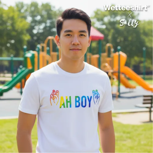 Singapore streetwear — man outdoors at playground wearing Wetteeshirt x Solly white graphic t-shirt with rainbow ‘AH BOY’ text and colorful hand symbols, casual lifestyle fashion shot with slides and climbing structures in sunny park background.