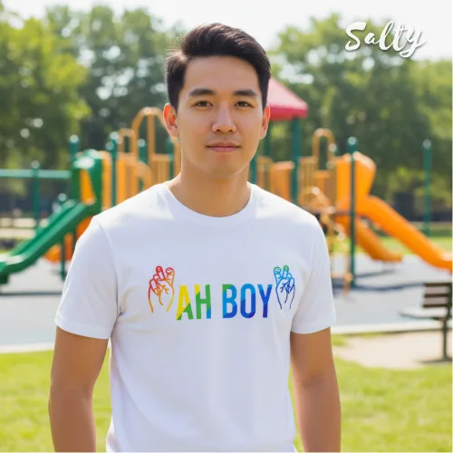 Singapore streetwear — man outdoors at playground wearing Wetteeshirt x Solly white graphic t-shirt with rainbow ‘AH BOY’ text and colorful hand symbols, casual lifestyle fashion shot with slides and climbing structures in sunny park background.