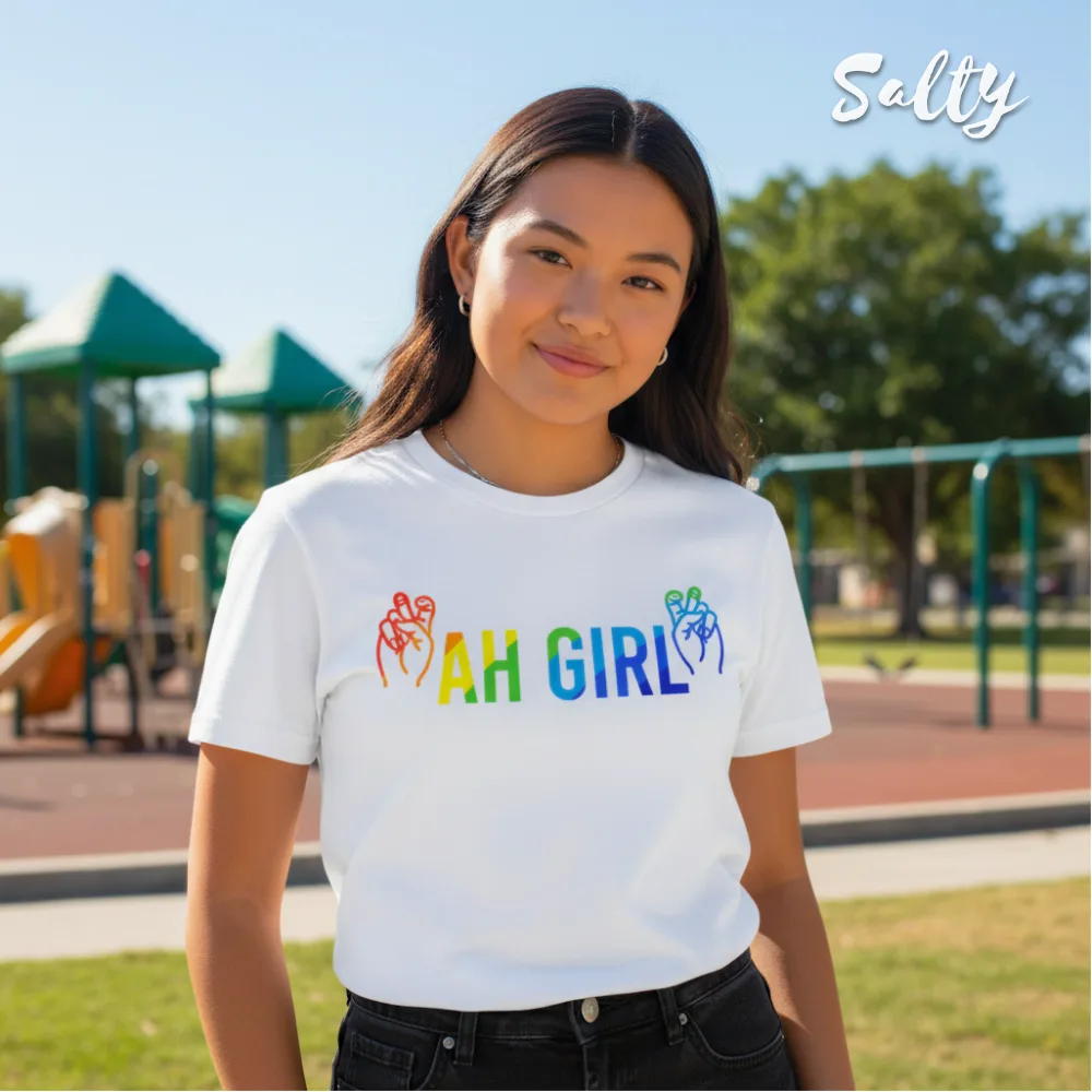 Singapore streetwear — person outdoors at playground wearing Wetteeshirt x Salty white graphic t-shirt with rainbow ‘AH GIRL’ text and red/blue hand gesture illustrations, casual lifestyle fashion shot with colorful play structures in background.