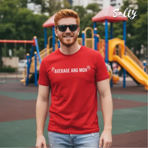 Outdoor streetwear photo of a bearded man in sunglasses wearing a bright red graphic tee with bold white text ‘AVERAGE ANG MOH,’ standing in front of a colorful Singapore playground, part of the Wet Tee Shirt® x Salty local culture collection.