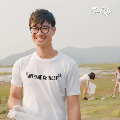 Outdoor photo of a man in glasses wearing a white graphic tee with bold text ‘AVERAGE CHINESE’ and small side graphics, standing on grass near water and hills during a cleanup activity, part of the Wet Tee Shirt® x Salty Singapore streetwear collection.