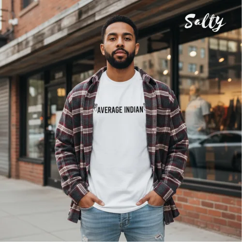 Urban streetwear photo of a man in glasses wearing a white graphic tee with bold black text ‘AVERAGE INDIAN,’ styled with an open plaid shirt and light blue jeans, standing on a sidewalk in front of a brick building with clothing displays, part of the Wet Tee Shirt® x Salty Singapore collection.