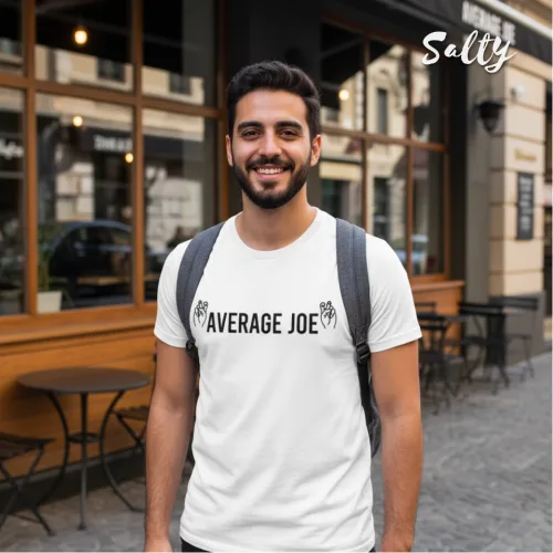 Urban streetwear photo of a man with gray backpack wearing a white graphic tee with bold black text ‘AVERAGE JOE’ and leaf motifs, standing on a cobblestone street near a café with wooden tables, part of the Wet Tee Shirt® x Salty Singapore collection.