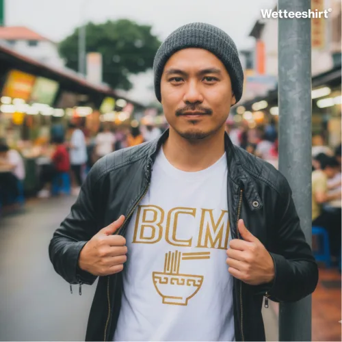 Person in gray beanie and black leather jacket at lively food street market, holding jacket open to show white Wetteeshirt® BCM tee with gold noodle bowl and chopsticks design, casual unisex Singapore streetwear inspired by hawker culture.