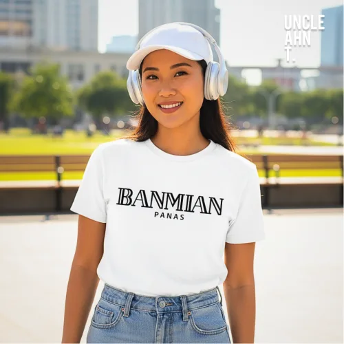 Casual lifestyle photo of a man in white cap and silver headphones wearing a white graphic tee with bold black text ‘BANMIAN PANAS,’ standing in an urban park with greenery and buildings, part of Wet Tee Shirt® x Uncle Ahn T Singapore streetwear collection.