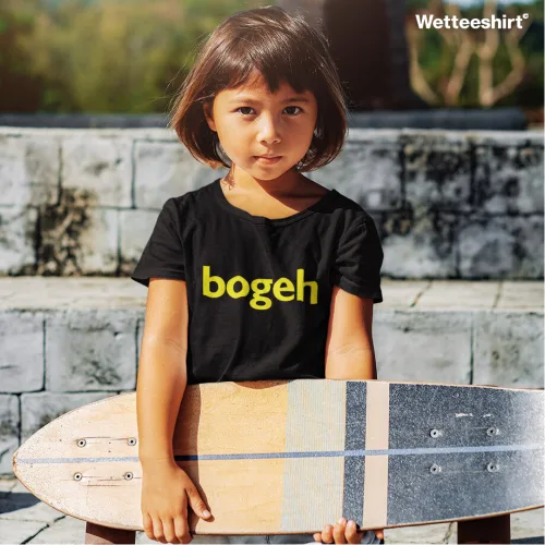 Child outdoors holding longboard skateboard, wearing black Wetteeshirt® Bogeh tee with bold yellow text design, casual kids streetwear fashion in sunny setting.jpg