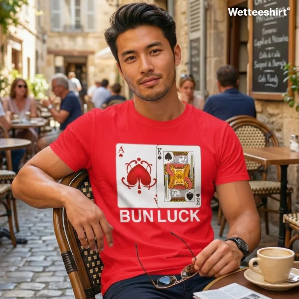 Man at outdoor café on cobblestone street wearing bright red Wetteeshirt® Bun Luck tee with playful Ace of Hearts and King of Clubs card designs featuring bun illustrations and bold white ‘BUN LUCK’ text, quirky unisex streetwear fashion.jpg