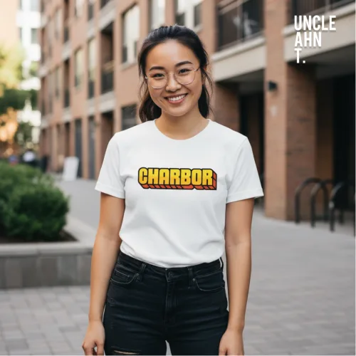 Urban streetwear photo of a man in glasses and dark shorts wearing a white graphic tee with bold yellow‑orange gradient text ‘CHARBOR’ outlined in black, standing on a paved walkway with brick buildings in the background, part of Wet Tee Shirt® x Uncle Ahn T Singapore collection.