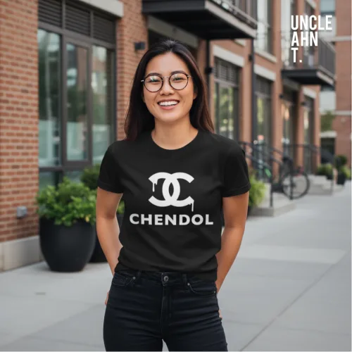 Urban streetwear photo of a man in glasses and dark pants wearing a black parody graphic tee with bold white ‘CHENDOL’ text beneath a double‑C logo design, standing on a sidewalk with brick buildings and large windows, part of Wet Tee Shirt® x Uncle Ahn T Singapore collection.