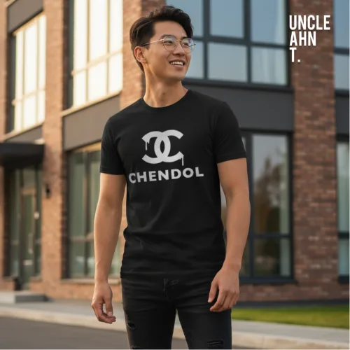 Streetwear photo of a man in a black parody graphic tee with bold white ‘CHENDOL’ text beneath a stylized double‑C logo, standing outside a modern building with glass windows and brick accents, part of Wet Tee Shirt® x Uncle Ahn T Singapore collection.