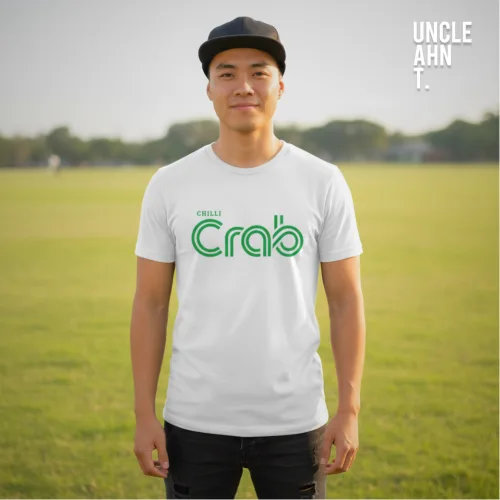 "Casual outdoor photo of a man in a black cap and dark pants wearing a white graphic tee with bold green stylized text ‘CRAB’ and smaller text ‘CHALL’ above it, standing on a grassy field with trees in the background, part of Wet Tee Shirt® x Uncle Ahn T Singapore streetwear collection.