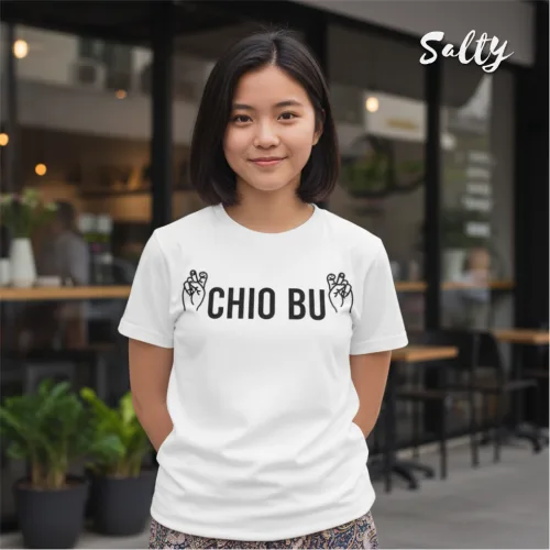 Lifestyle streetwear photo of a woman wearing a white graphic tee with bold black text ‘CHIO BU’ and hand illustrations, standing outside a café with glass windows and potted plants, part of the Wet Tee Shirt® x Salty Singapore collection.