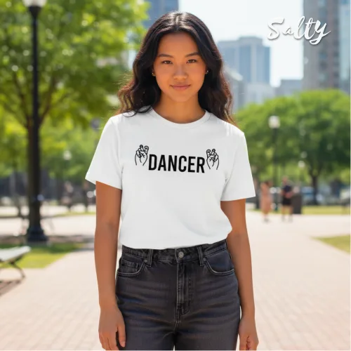 Singapore streetwear — person outdoors on paved park pathway wearing Wetteeshirt® Salty white graphic t-shirt with bold black ‘DANCER’ text flanked by dancing figure illustrations, styled with high‑waisted dark jeans, casual lifestyle fashion shot with trees and cityscape in background.