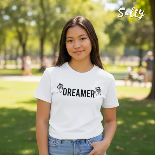 Singapore streetwear — young person outdoors in park wearing Wetteeshirt® x Solly white graphic t-shirt with bold black ‘DREAMER’ text flanked by peace sign hand illustrations, styled with light blue jeans, casual lifestyle fashion shot with trees, grass, and people in background.