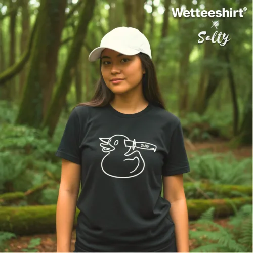 Singapore streetwear — person in forest wearing Wetteeshirt® X Salty black graphic t-shirt with bold duck illustration stabbed by knife labeled ‘SALTY,’ styled with white cap, lifestyle fashion shot with tall trees and sunlight background.