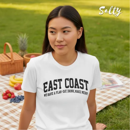 Singapore streetwear — young woman outdoors on picnic blanket wearing Wetteeshirt × Salty white graphic t-shirt with bold black text ‘EAST COAST WE HAVE A PLAN: EAT, DRINK, MAKE MERRY,’ casual lifestyle fashion shot with picnic basket, fruits, and grassy park background.