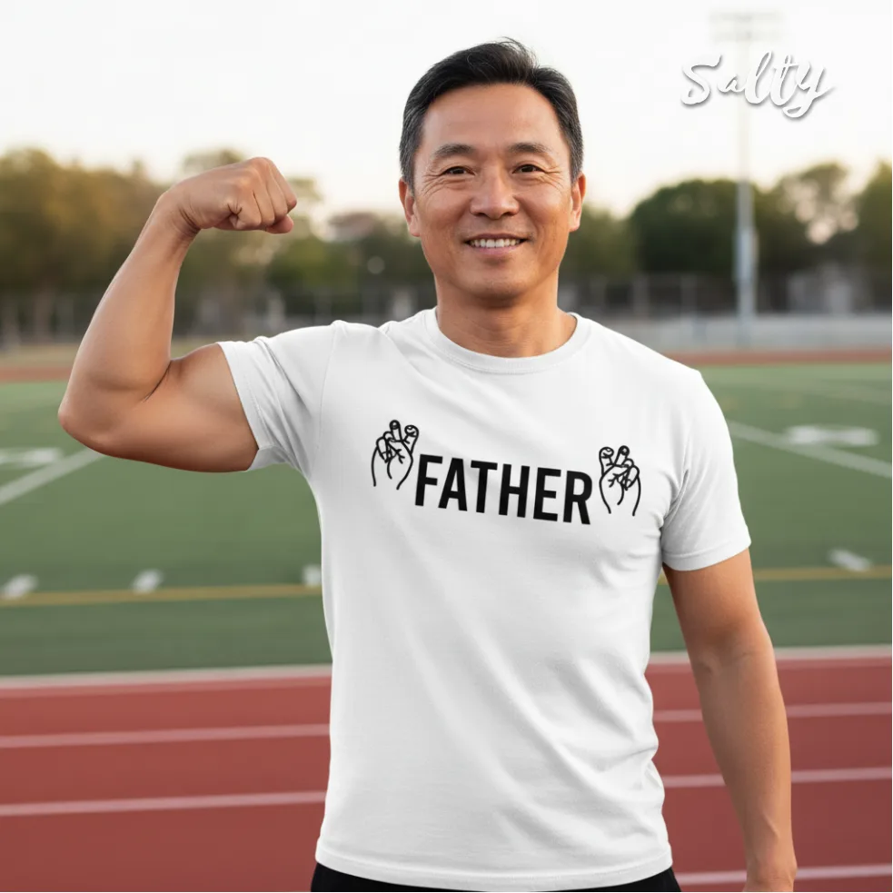 "Outdoor lifestyle photo of a smiling man flexing his arm on a running track, wearing a white graphic tee with bold black text ‘FATHER’ and fist‑bump design, set against a green athletic field, part of the Wet Tee Shirt® x Salty Singapore streetwear collection.