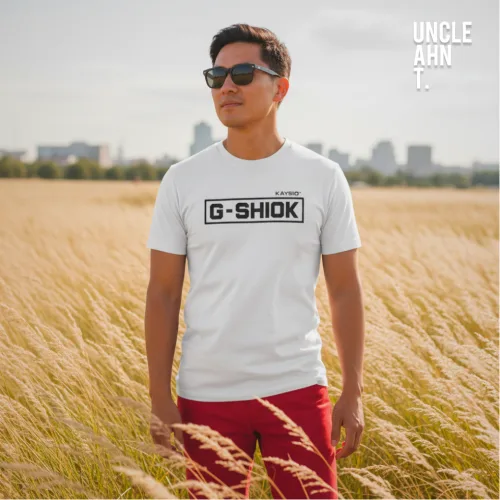 "Singapore streetwear EAT MORE G‑SHIOK white short sleeve T-shirt, bold black text in rectangular border, casual unisex tee by Uncle Ahn T, premium cotton playful design photographed outdoors in golden grass with city skyline