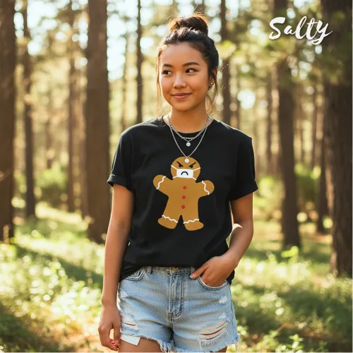 "Singapore streetwear — person in sunlit forest wearing Wetteeshirt® X Salty black graphic t-shirt featuring angry gingerbread man with red buttons and white icing details, styled with light blue denim shorts and layered necklaces, lifestyle fashion shot with tall trees and greenery.