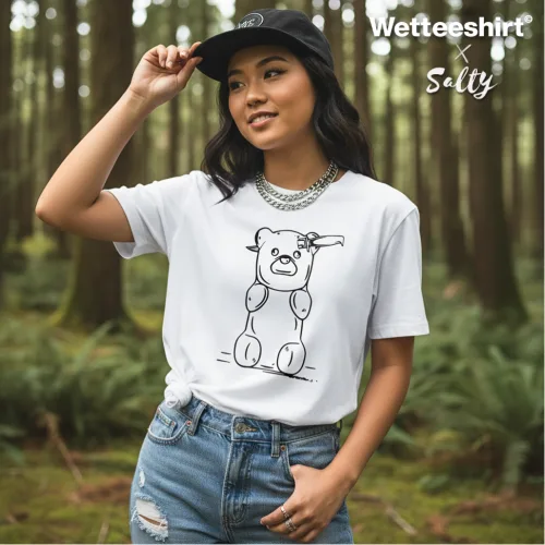 Singapore streetwear — person in forest wearing Wetteeshirt X Salty white graphic t-shirt with playful cartoon bear made of balloon segments waving, styled with ripped blue jeans, black cap, and silver chain necklace, lifestyle fashion shot with tall trees and greenery.