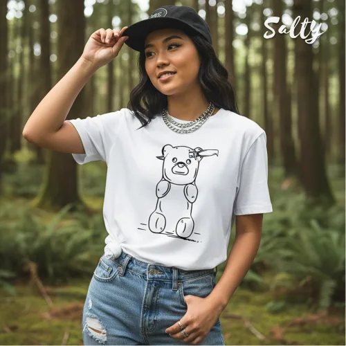 Singapore streetwear — person in forest wearing Wetteeshirt X Salty white graphic t-shirt with playful cartoon bear made of balloon segments waving, styled with ripped blue jeans, black cap, and silver chain necklace, lifestyle fashion shot with tall trees and greenery.