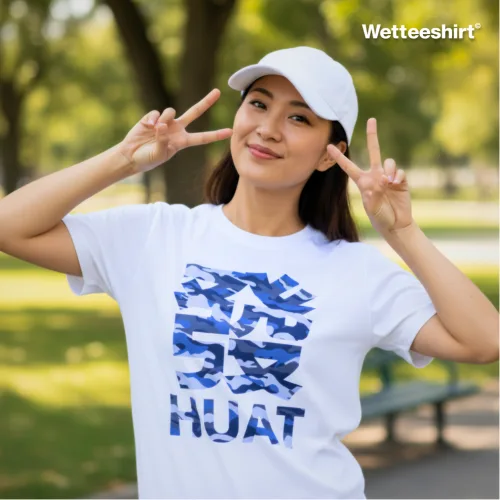 "White casual T-shirt with bold blue camouflage Chinese character ‘發’ and ‘HUAT’ print, styled with white cap and double peace sign gesture, worn outdoors in park setting — cheerful streetwear fashion celebrating prosperity and good fortune.