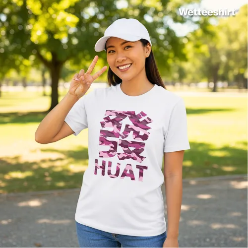 "White casual T-shirt with bold camouflage Chinese character and ‘HUAT’ print, styled with white cap and peace sign gesture, worn outdoors in sunny park setting with trees — cheerful streetwear fashion celebrating prosperity and good fortune.