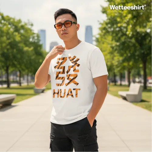 White casual T-shirt with bold gold-pattern Chinese character and large ‘HUAT’ print, styled with sunglasses and black shorts, worn outdoors on paved pathway with trees and tall buildings — modern streetwear fashion celebrating prosperity and good fortune.