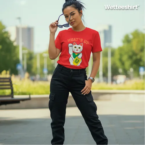 Red streetwear T-shirt featuring playful ‘HUAT’S UP’ slogan with waving lucky cat graphic holding gold coin, styled with black cargo pants, wristwatch, and sunglasses in an urban park setting — trendy fashion blending festive humor with East Asian prosperity symbolism.