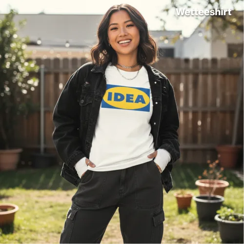 White casual T-shirt with bold yellow ‘IDEA’ logo inside blue oval, styled with black denim jacket and dark cargo pants, worn outdoors in backyard with wooden fence, potted plants, and house — witty streetwear parody inspired by IKEA design.