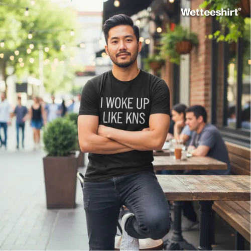 Black casual T-shirt with bold white ‘I WOKE UP LIKE KNS’ slogan, styled with denim jacket, layered silver necklaces, and urban street backdrop with café tables and pedestrians — witty streetwear fashion blending humor with casual city lifestyle.