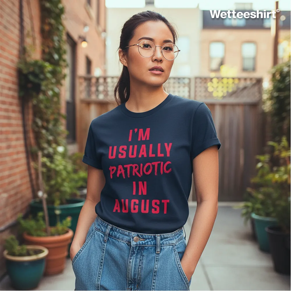 Navy blue casual T-shirt with bold red ‘I’M USUALLY PATRIOTIC IN AUGUST’ slogan, styled with glasses and high-waisted jeans, worn outdoors in urban courtyard with brick buildings and potted plants — witty streetwear fashion with playful seasonal humor.