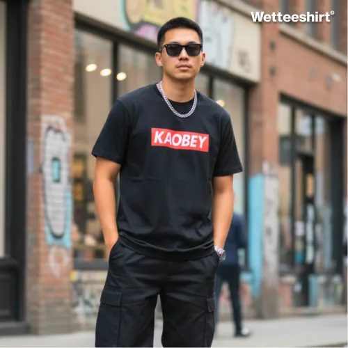 Black streetwear T-shirt with bold white ‘KAOBEY’ text on red rectangular background, styled with sunglasses, chain necklace, watch, and black cargo pants, worn outdoors on graffiti-covered city street — edgy urban fashion parody inspired by Supreme logo style.