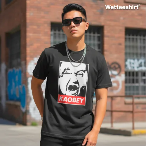 Black streetwear T-shirt with bold graphic face design and white ‘KAOBEY’ text on red background, styled with sunglasses and silver chain necklace, worn outdoors in front of brick building with graffiti — edgy urban fashion parody inspired by iconic OBEY street art style.