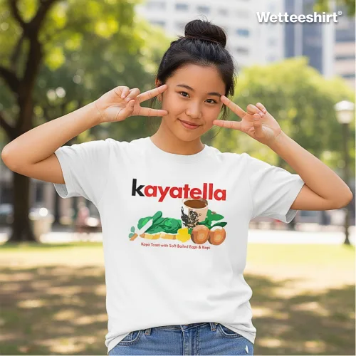 White casual T-shirt with bold red ‘kayatella’ text and playful cartoon cat surrounded by spinach, tomatoes, onions, and rice dish caption ‘Kaya Tomato with Soft Boiled Egg & Rice,’ styled outdoors in urban park with peace sign pose — quirky streetwear blending food humor with cute design.