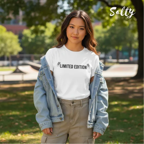 Singapore streetwear — person outdoors in park skate area wearing Wetteeshirt® x Salty white graphic t-shirt with bold black ‘LIMITED EDITION’ text flanked by hand graphics, styled with light blue denim jacket and beige cargo pants, casual lifestyle fashion shot with grass, trees, and skate park background.