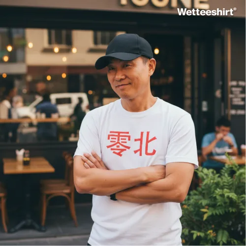 White casual T-shirt with bold red Chinese characters ‘零北,’ styled with black baseball cap and arms crossed, worn outdoors in front of café with glass windows and outdoor seating — modern streetwear fashion with cultural text design.