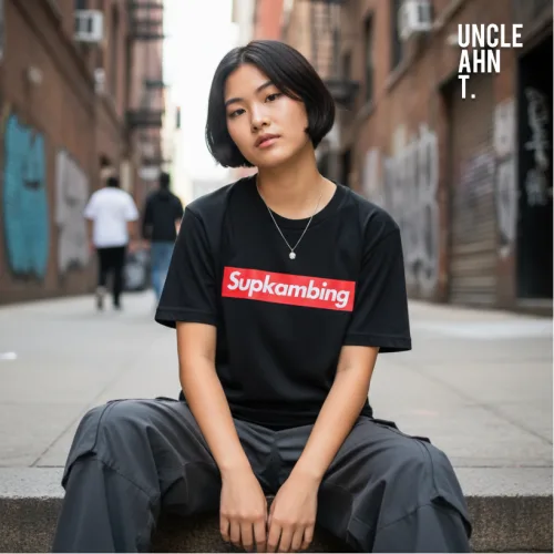 "Singapore streetwear Supkambing black short sleeve T-shirt, bold red rectangular logo with white text parody design, casual unisex tee by Uncle Ahn T, premium cotton playful design photographed outdoors in urban graffiti street setting