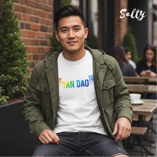 Casual urban photo of a man sitting at an outdoor café in a green jacket over a white graphic tee with rainbow text ‘YAN DAO,’ surrounded by brick walls, greenery, and other diners, part of the Wet Tee Shirt® x Salty Singapore streetwear collection.