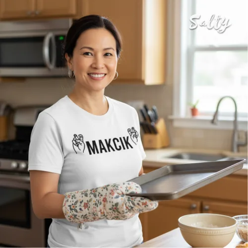 Cheerful kitchen photo of a woman in patterned oven mitts holding a baking tray, wearing a white graphic tee with bold black text ‘MAKCIK’ and side figures, surrounded by wooden cabinets and natural light, part of the Wet Tee Shirt® x Salty Singapore streetwear collection.
