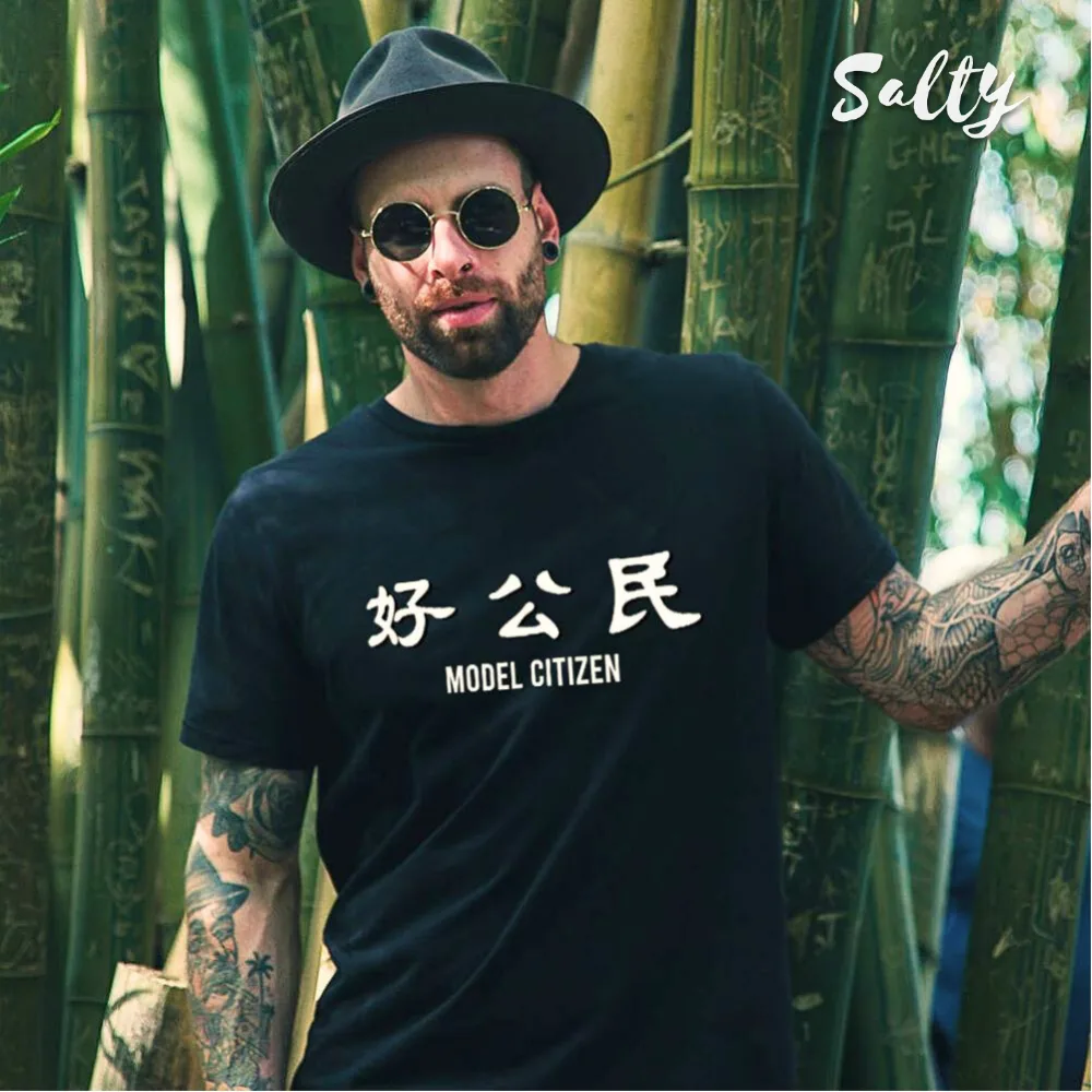 Black graphic t-shirt with bilingual text 好公民 (Good Citizen) and MODEL CITIZEN in bold white lettering. Styled with black hat, round sunglasses, tattoos, and beard against bamboo backdrop. Wet Tee Shirt® x Salty collaboration blending cultural design with edgy streetwear fashion.