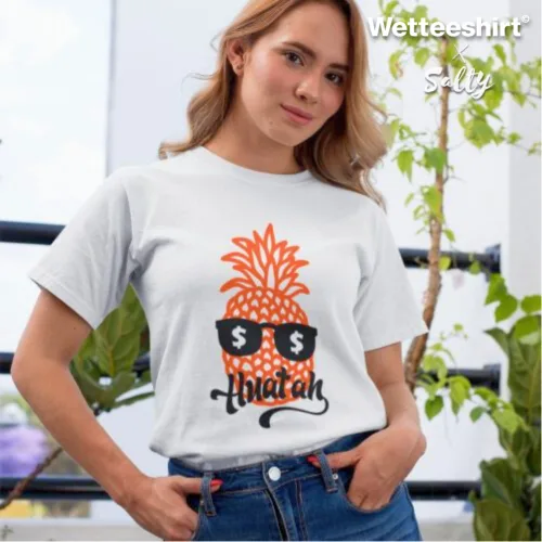 Singapore streetwear — person indoors wearing Wetteeshirt® Silly white graphic t-shirt with playful orange pineapple in black sunglasses featuring dollar sign lenses, stylized ‘Aloha ah’ text below, lifestyle fashion shot with green plants in bright background.