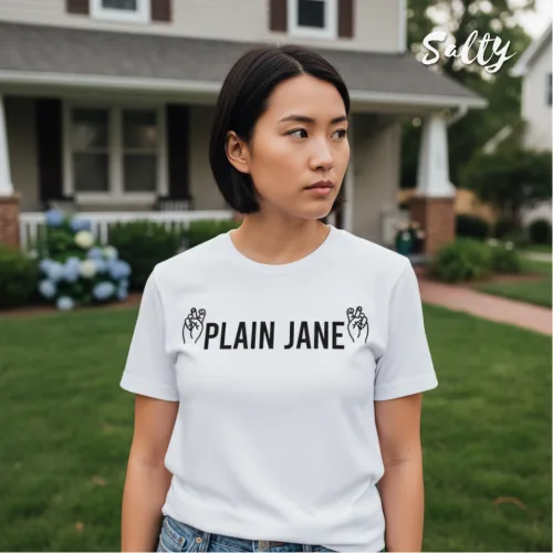 Lifestyle streetwear photo of a woman wearing a white graphic tee with bold black text ‘PLAIN JANE’ and hand illustrations, standing in front of a suburban house with lawn and flowers, part of the Wet Tee Shirt® x Salty Singapore collection.