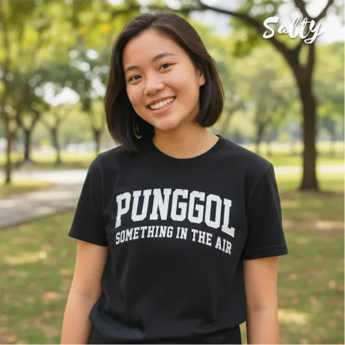 Singapore streetwear — smiling person outdoors in park wearing Wetteeshirt® Salty black graphic t-shirt with bold white ‘PUNGGOL SOMETHING IN THE AIR’ text, casual lifestyle fashion shot with trees and pathway in background.