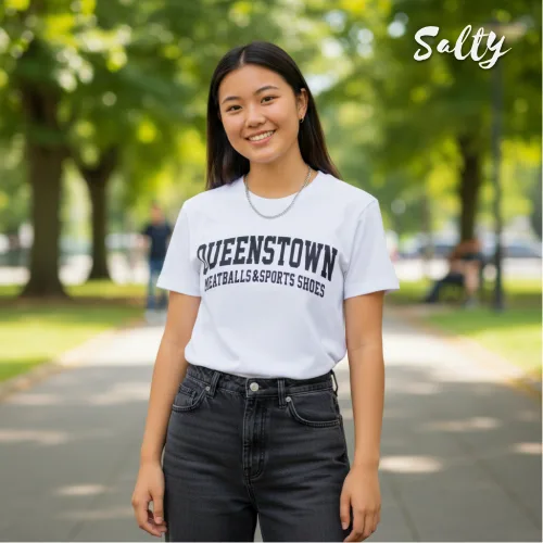 Singapore streetwear — person outdoors on tree‑lined pathway wearing Wetteeshirt® × Salty white graphic t-shirt with bold black ‘QUEENSTOWN NETBALLS & SPORTS SHOES’ text, styled with dark jeans, casual lifestyle fashion shot with sunlight filtering through park trees.