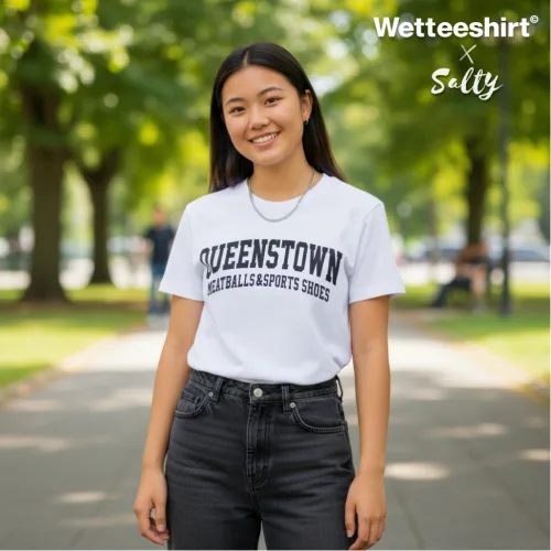 Singapore streetwear — person outdoors on tree‑lined pathway wearing Wetteeshirt® × Salty white graphic t-shirt with bold black ‘QUEENSTOWN NETBALLS & SPORTS SHOES’ text, styled with dark jeans, casual lifestyle fashion shot with sunlight filtering through park trees."