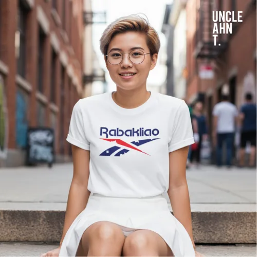 Singapore streetwear Rabakliao white short sleeve T-shirt, bold blue and red stylized text with swoosh graphic, casual unisex tee by Uncle Ahn T, premium cotton playful design photographed outdoors in urban alley