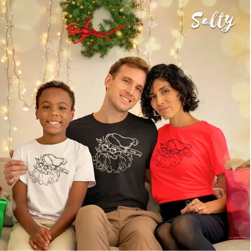 Christmas family photo with matching Santa Claus face graphic t-shirts in black, white, and red. Cozy holiday setting with string lights, wreath, and wrapped gifts. Wet Tee Shirt® x Salty collaboration showcasing festive apparel for family celebrations and holiday streetwear fashion.