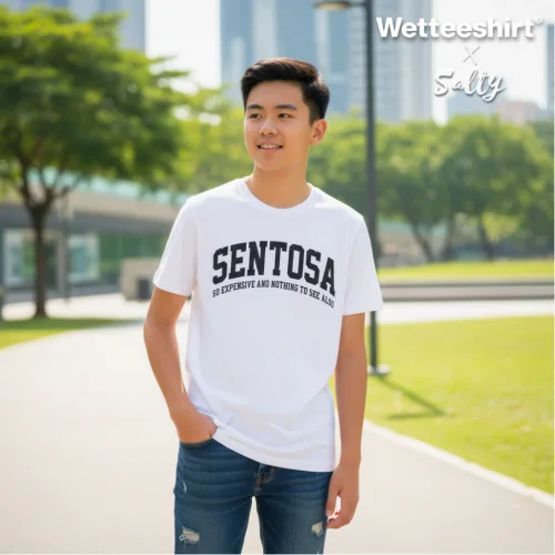 Singapore streetwear — young person outdoors in park with cityscape background wearing Wetteeshirt x 5aily white graphic t-shirt with bold black ‘SENTOSA’ text and smaller slogan ‘An Experience Like Nothing Else On Earth,’ styled with blue jeans, casual lifestyle fashion shot on sunny day.