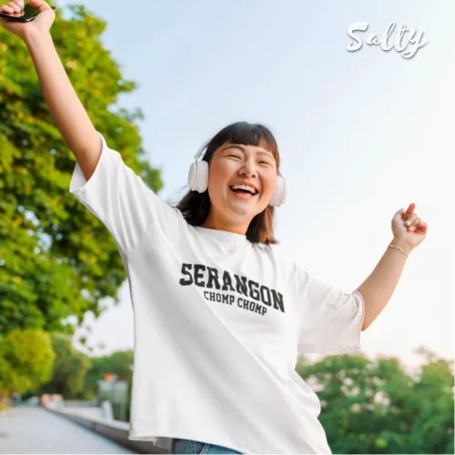 Singapore streetwear — joyful person outdoors on tree‑lined pathway wearing Wetteeshirt x Salty white graphic t-shirt with bold black ‘SERANGOON CHOMP CHOMP’ text, smiling with arms raised and white headphones, casual lifestyle fashion shot with greenery and clear sky background.